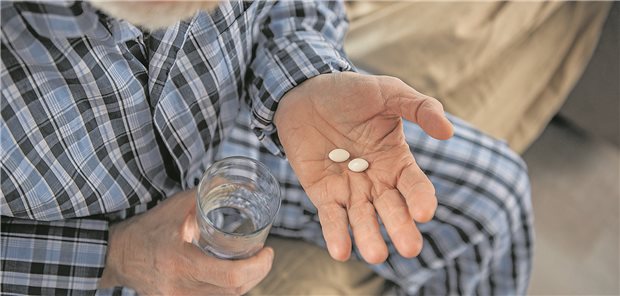 Ein Mann sitzt auf seinem Bett. In der Hand hat er ein Glas Wasser und Medikamente für seinen Blutdruck.