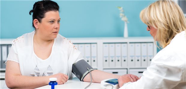 Ärztin misst den Blutdruck einer Patientin. Ihr Arm liegt auf dem Tisch, die Manschette ist am Oberarm befestigt.