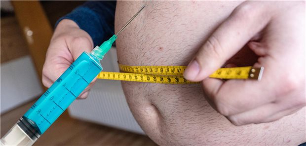 PKV muss Abnehmspritze nur bei Einbettung in Therapiekonzept zahlen Ein Mann hält eine große Spritze, während er mit einem Maßband den Umfang des Bauch misst.