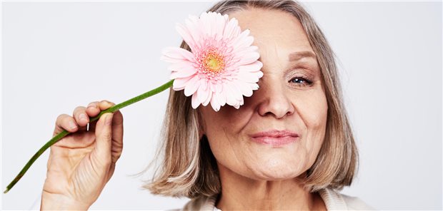 Frau mittleren Alters hält sich eine rosa Blume vor das Gesicht und lächelt.