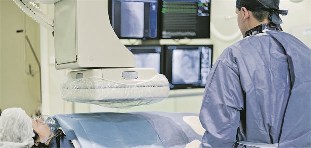 Studie: Krankenhausübernahmen senken Sterblichkeit nach Herzinfarkten Das Bild zeigt einen Blick in ein Herzkatheterlabor. Ein Patient wird mit bildgebenden Instrumenten untersucht. Ein Arzt betrachtet die Ergebnisse auf dem Bildschirm.