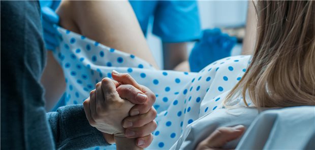 Wie sich Traumatisierung und Retraumatisierung während der Geburt vermeiden lassen Nahaufnahme einer Frau bei der Geburt, während ihr Ehemann ihr zur Unterstützung die Hand hält und ein Geburtshelfer bei der Entbindung assistiert.