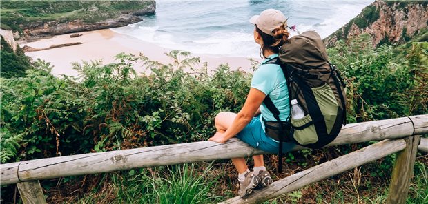Reisemedizin: Wie Sie die Beratung von jüngeren Patienten angehen können Eine junge Backpackerin sitzt bei einer Klippe und schaut auf einen Strand herunter.