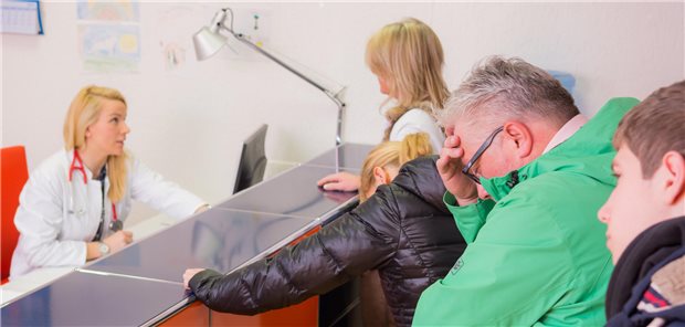 Hohe Arbeitsbelastung der MFA: Welche Strategien zur Entlastung sich lohnen Eine Ärztin sitzt hinter dem Anmeldepult. Es warten viele Patientinnnen und Patienten in einer Schlange.