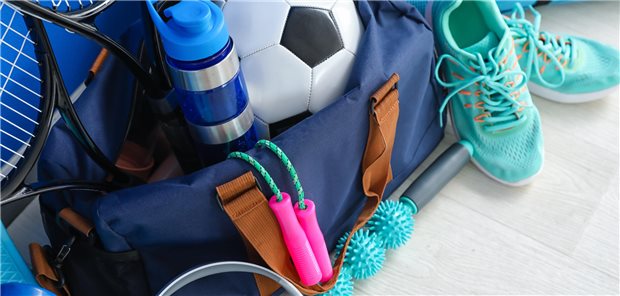 Mit einer Stunde mehr Sport die Brustkrebsmortalität senken? Eine Sporttasche liegt am Boden und ist gefüllt mit Sportsachen, wie einem Springseil, einem Fußball, Laufschuhen und Tennisschlägern.