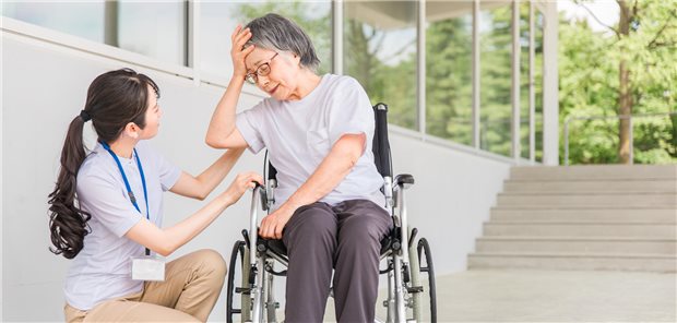 Diese Auswirkungen hat Hitze auf Menschen mit neurologischen Erkrankungen Eine ältere Frau im Rollstuhl fasst sich mit der Hand an den Kopf. Eine Pflegerin sitzt daneben und kümmert sich.