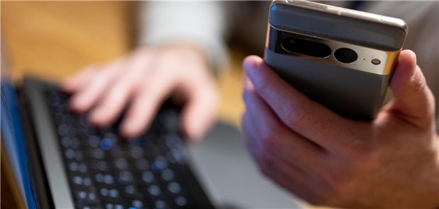 Eine Person sitzt am Computer und hält ihr Smartphone in der Hand.