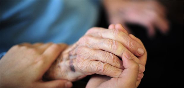 Unser Pflegehaus steht in Flammen Eine Pflegebegutachterin während eines Gesprächs mit einem älteren Mann, der mit einem Gehstock an einem Tisch sitzt.