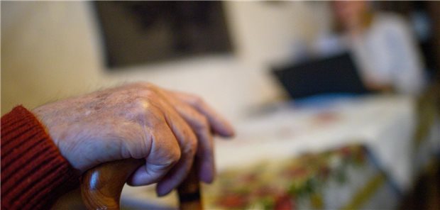 Verbände und Kassen zu Warkens Pflegeplänen: „Es besteht weiter erheblicher Nachbesserungsbedarf“ Die Hand einer pflegebedürftigen Seniorin an einem Gehstock; im Hintergrund einer Fachkraft mit Laptop.