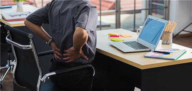 Rückenschmerzen: „Falsche Diagnosen lassen viele Therapien scheitern!“ Ein Mann sitzt am Schreibtisch und hält sich die Hände an den unteren Rücken.