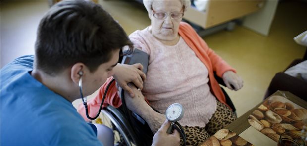 Heilkundeübertragung: Wo Substitution die ambulante Versorgung verbessern könnte Eine männliche Pflegekraft misst Blutdruck bei einer älteren Frau im Rollstuhl.