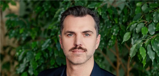 Digitales Gesundheitswesen? „Aktuell befinden wir uns noch in der Phase der Elektrifizierung“ Manuel Nothelfer steht vor einer Blätterwand und schaut in die Kamera.