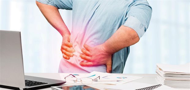 Warum Praxen bei Patienten mit leichten Beschwerden an die Rentenversicherung denken sollten Arbeitnehmer steht mit seinem Rücken zu seinem Schreibtisch auf dem ein Laptop steht und hält sich aufgrund von Rückenschmerzen seinen Rücken.